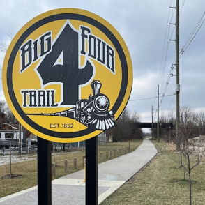 Sign for "Big Four Trail" features a steam locomotive illustration, set against a paved pathway and overcast sky, evoking adventure.