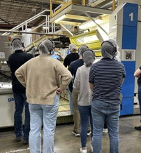 Group of people in hairnets engage around an industrial machine, showcasing curiosity, collaboration, and professionalism.
