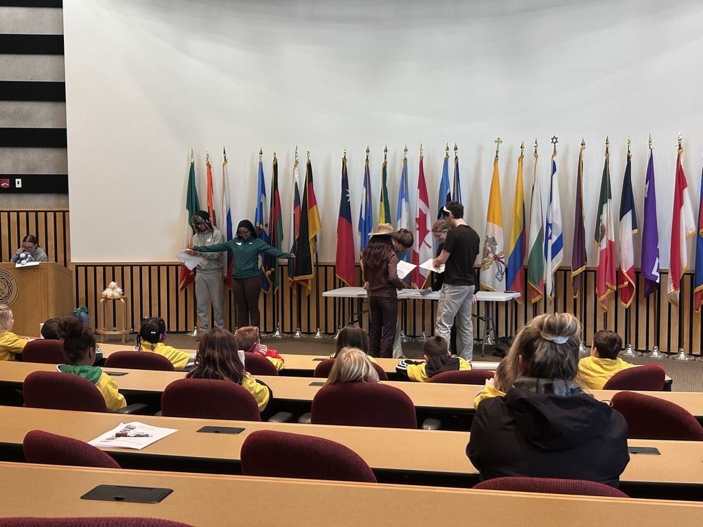 A lively auditorium scene shows presenters and engaged children in yellow jackets, with international flags symbolizing collaboration.