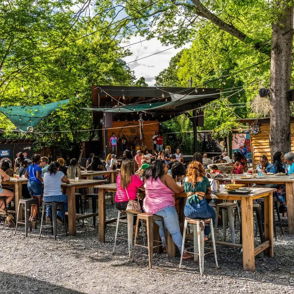 Outdoor gathering with lively patrons at wooden tables, enjoying food and live entertainment in a sunny, vibrant atmosphere.
