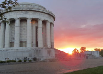 A grand monument with classical elements stands under a vibrant sunset, surrounded by trees and a tranquil pathway.