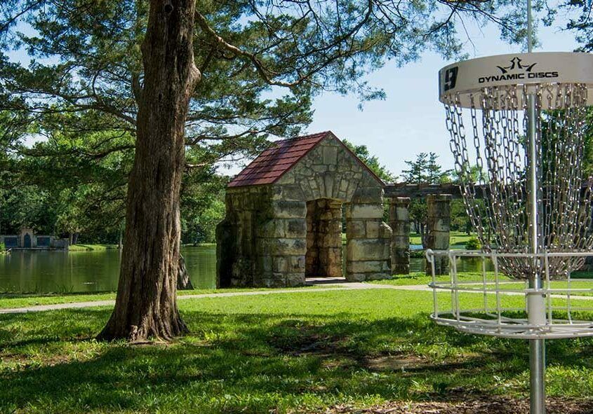 Serene outdoor scene with a disc golf basket, a stone structure, calm water, and towering trees, evoking tranquility and leisure.