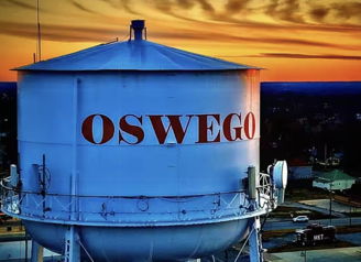 Water tower with "OSWEGO" in red letters against a vibrant sunset sky, evoking nostalgia and community warmth.