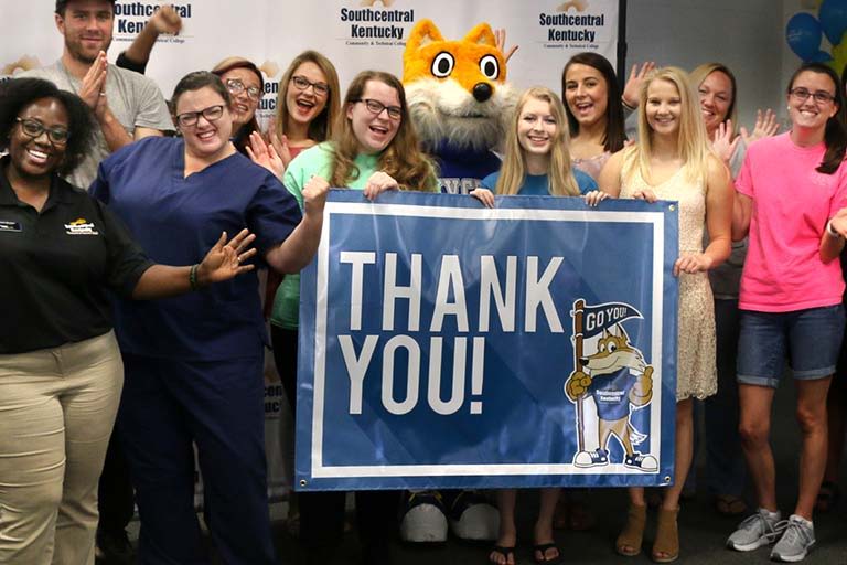 Group celebration with mascot, smiling people, and a "THANK YOU!" banner, conveying joy, appreciation, and community spirit.
