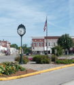 Charming town square with vibrant flowers, brick buildings, a clock, and an American flag, evoking community and nostalgia.
