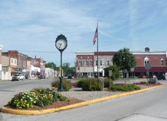 Charming town square with vibrant flowers, brick buildings, a clock, and an American flag, evoking community and nostalgia.
