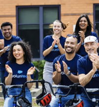 A diverse group celebrates outdoors in blue t-shirts, smiling and clapping, conveying unity, joy, and community spirit.