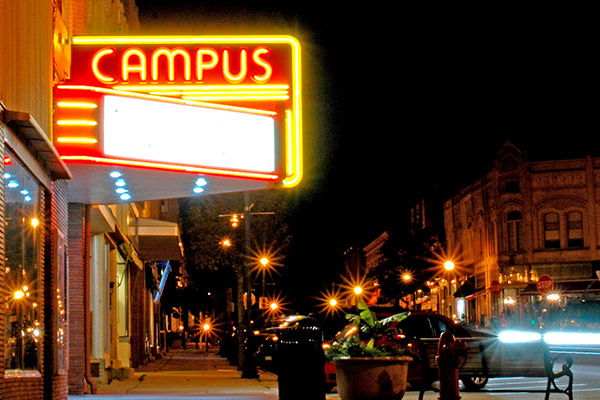 Vibrant nighttime street scene with a retro neon "CAMPUS" sign, evoking nostalgia, warmth, and a lively community atmosphere.