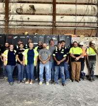 A group in safety gear stands together in a workshop, showcasing teamwork, pride, and camaraderie in a hardworking atmosphere.