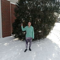 Person in a light green sweatshirt poses playfully in front of a snowy evergreen, evoking joy and winter cheer.