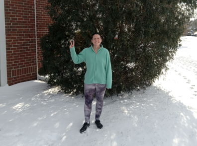 Person in a light green sweatshirt poses playfully in front of a snowy evergreen, evoking joy and winter cheer.