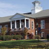 A stately brick building with columns and a cupola, set in vibrant, well-landscaped surroundings on a clear day.