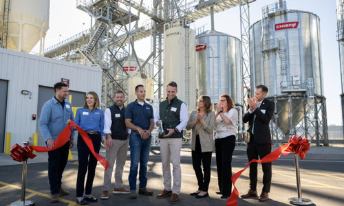 Group smiles at a ribbon-cutting ceremony for a new facility, with silos in the background, highlighting community and celebration.