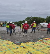 Construction site with workers pouring concrete, wearing safety gear, showcasing teamwork and dedication under an overcast sky.