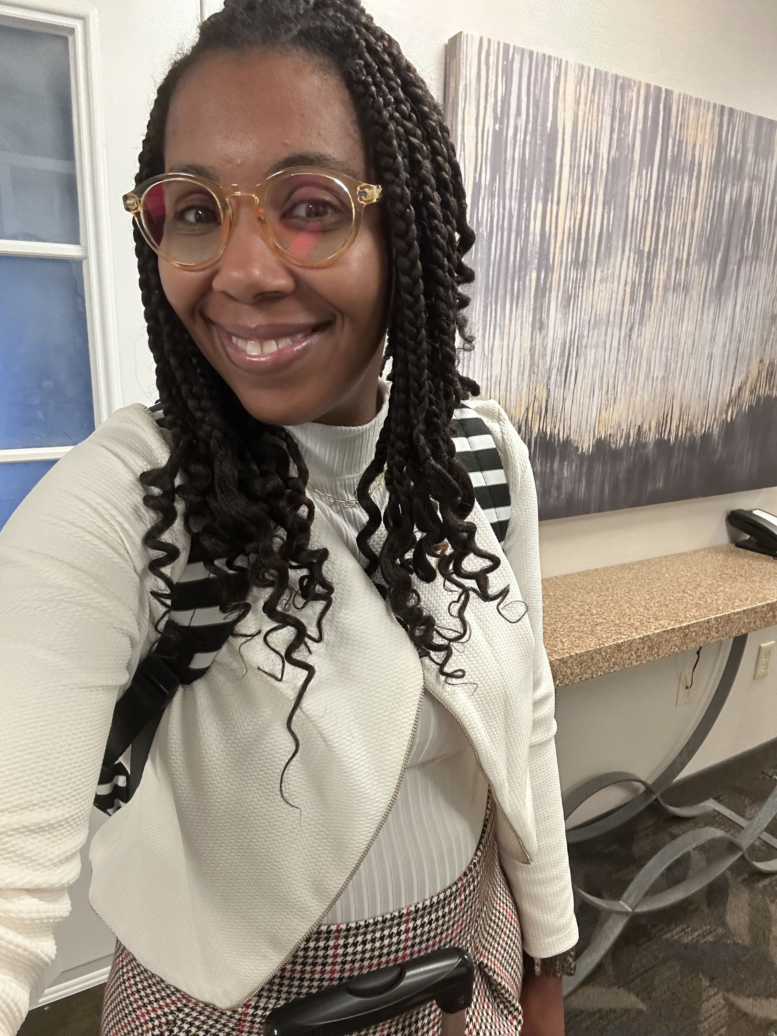 Person with braided hair and stylish glasses wears a fashionable outfit in a modern indoor space, exuding happiness and confidence.