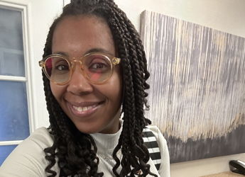 Person with braided hair and stylish glasses wears a fashionable outfit in a modern indoor space, exuding happiness and confidence.