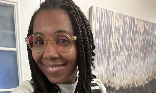 Person with braided hair and stylish glasses wears a fashionable outfit in a modern indoor space, exuding happiness and confidence.