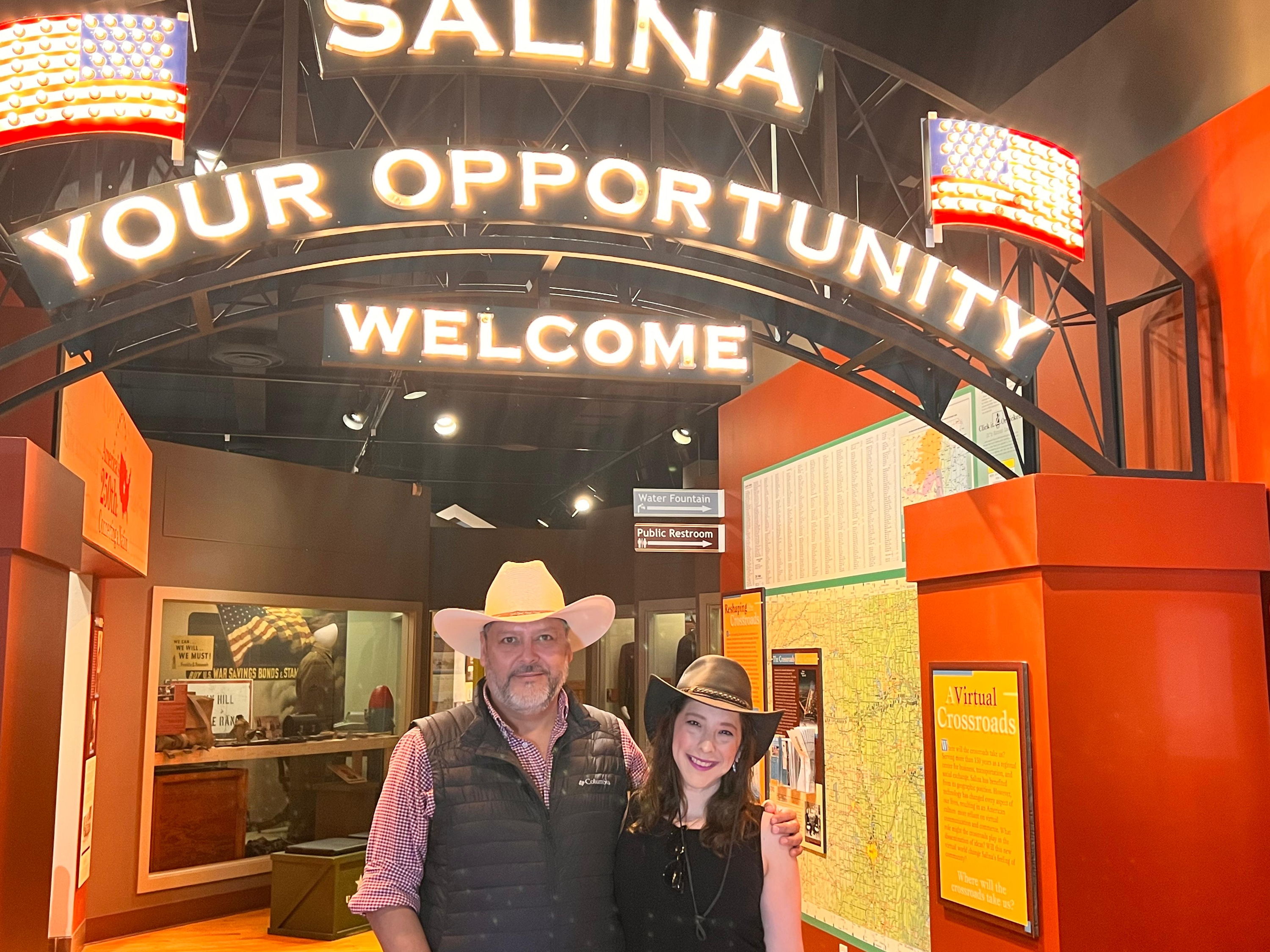 Two individuals stand under a festive arch reading "SALINA YOUR OPPORTUNITY WELCOME," radiating warmth and camaraderie.