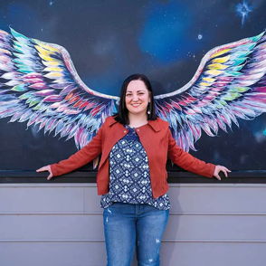 A smiling woman in casual attire stands before a colorful wing mural, creating a joyful and uplifting atmosphere.