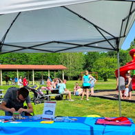 A sunny park scene features a tented table with items, families, and a colorful playground, evoking joy and community.