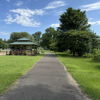 A tranquil pathway through green grass and trees, leading to a distant gazebo under a blue sky with clouds.