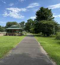 A tranquil pathway through green grass and trees, leading to a distant gazebo under a blue sky with clouds.