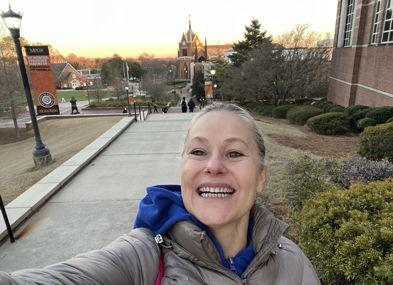 A person smiles while taking a selfie outdoors on campus, with a warm sunset creating a joyful and uplifting atmosphere.