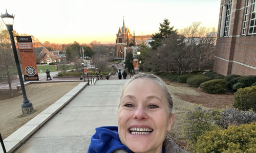 A person smiles while taking a selfie outdoors on campus, with a warm sunset creating a joyful and uplifting atmosphere.