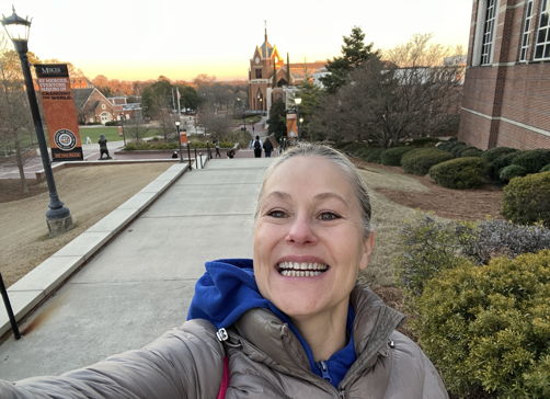 A person smiles while taking a selfie outdoors on campus, with a warm sunset creating a joyful and uplifting atmosphere.