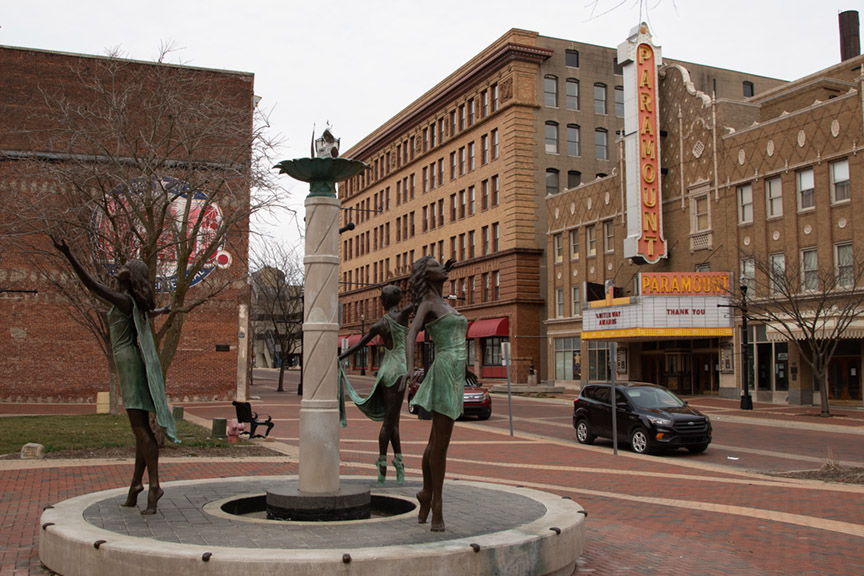 Plaza with a fountain and bronze sculptures of women, surrounded by historic buildings, evoking nostalgia and serenity.
