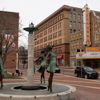 Plaza with a fountain and bronze sculptures of women, surrounded by historic buildings, evoking nostalgia and serenity.
