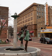 Plaza with a fountain and bronze sculptures of women, surrounded by historic buildings, evoking nostalgia and serenity.