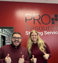 Two smiling individuals give a thumbs-up in front of a red wall displaying "PRO Resources Staffing Services," radiating positivity.
