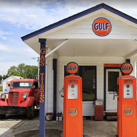 Vintage gas station with bright orange Gulf pumps, white and blue design, classic red trucks, evoking Americana nostalgia.