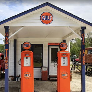 Vintage gas station with bright orange Gulf pumps, white and blue design, classic red trucks, evoking Americana nostalgia.