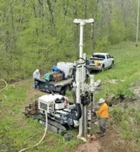 A drilling machine is operated by a person in orange gear, surrounded by greenery and work vehicles, showcasing industry in nature.