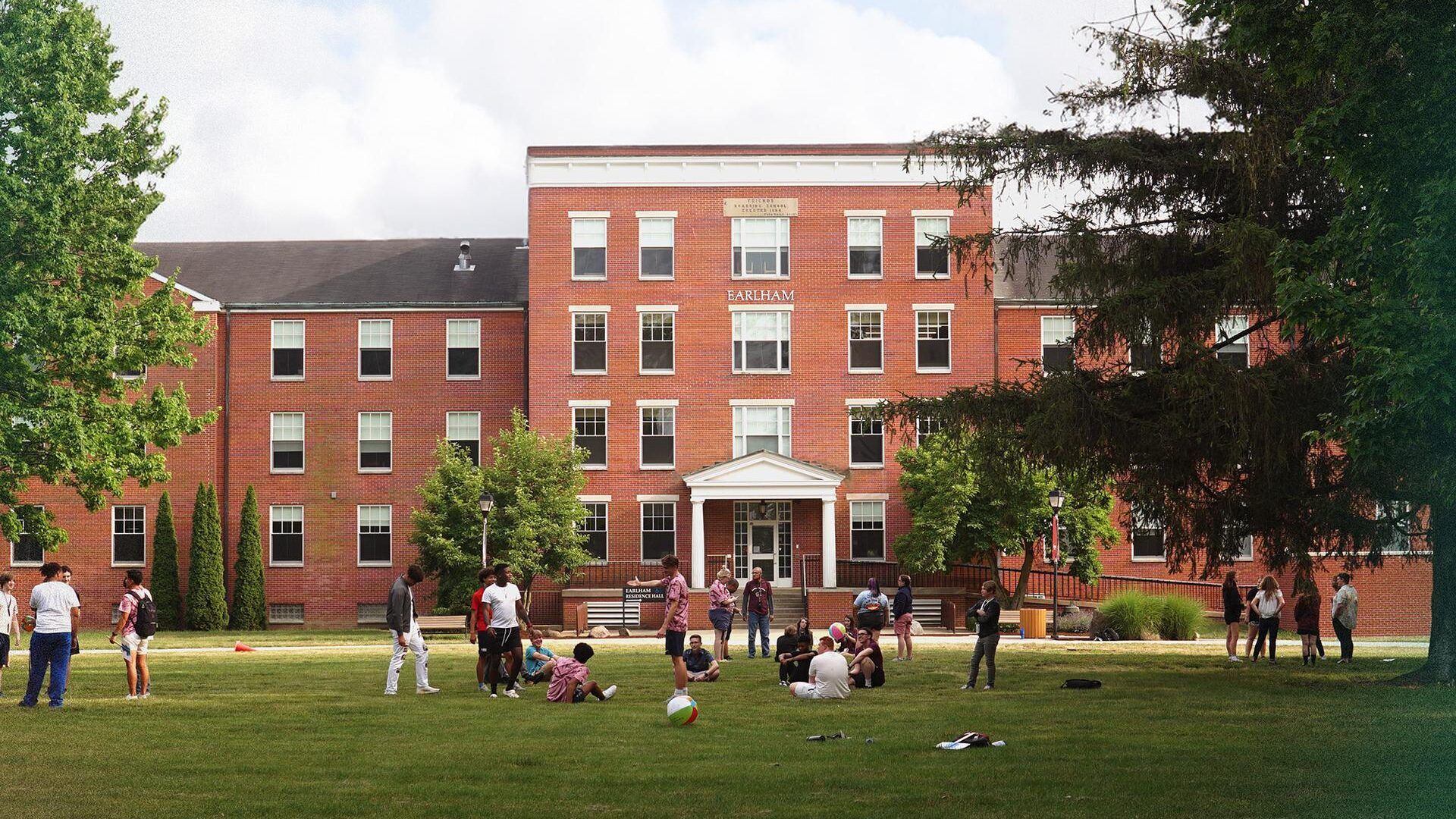A lively outdoor scene near a brick building, with children and young adults playing and socializing in a cheerful atmosphere.