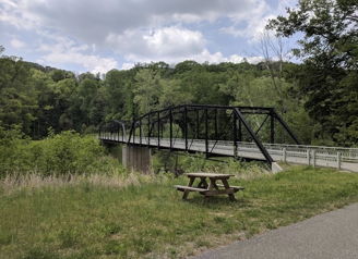 A classic truss bridge arches over a serene river, framed by lush greenery and a picnic table, evoking tranquility.