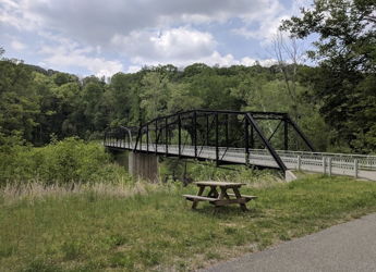 A classic truss bridge arches over a serene river, framed by lush greenery and a picnic table, evoking tranquility.