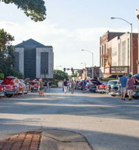 Vintage cars in various colors fill a lively street event, with people enjoying a charming community gathering under sunlight.