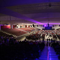 Indoor venue mostly empty, crowd gathered in front of a dimly lit stage, purple lighting creates an atmosphere of anticipation.