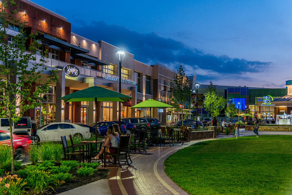 Twilight urban scene with shops, eateries, greenery, and warm lights, evoking relaxation and community vibes.