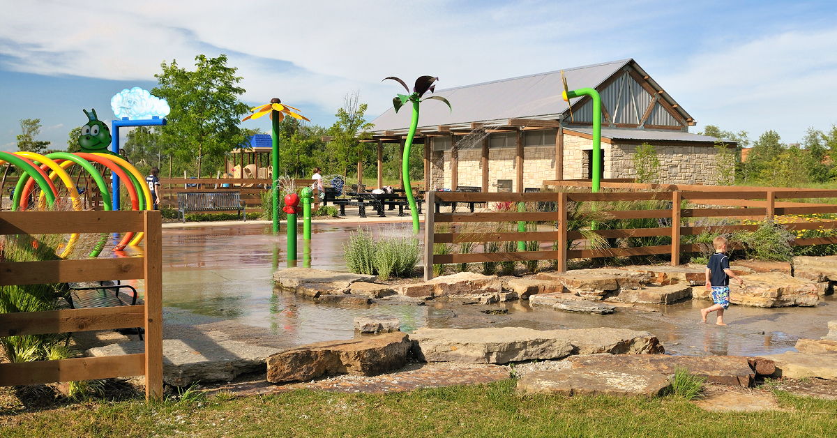 A child plays joyfully in a colorful water area in a sunny park, surrounded by greenery and a lively atmosphere.