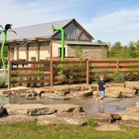 A child plays joyfully in a colorful water area in a sunny park, surrounded by greenery and a lively atmosphere.
