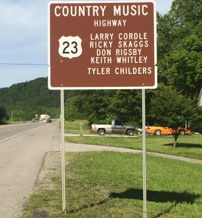 Sign for "Country Music Highway" 23, featuring notable artists, set in a lush, sunny rural backdrop, evoking nostalgia and joy.