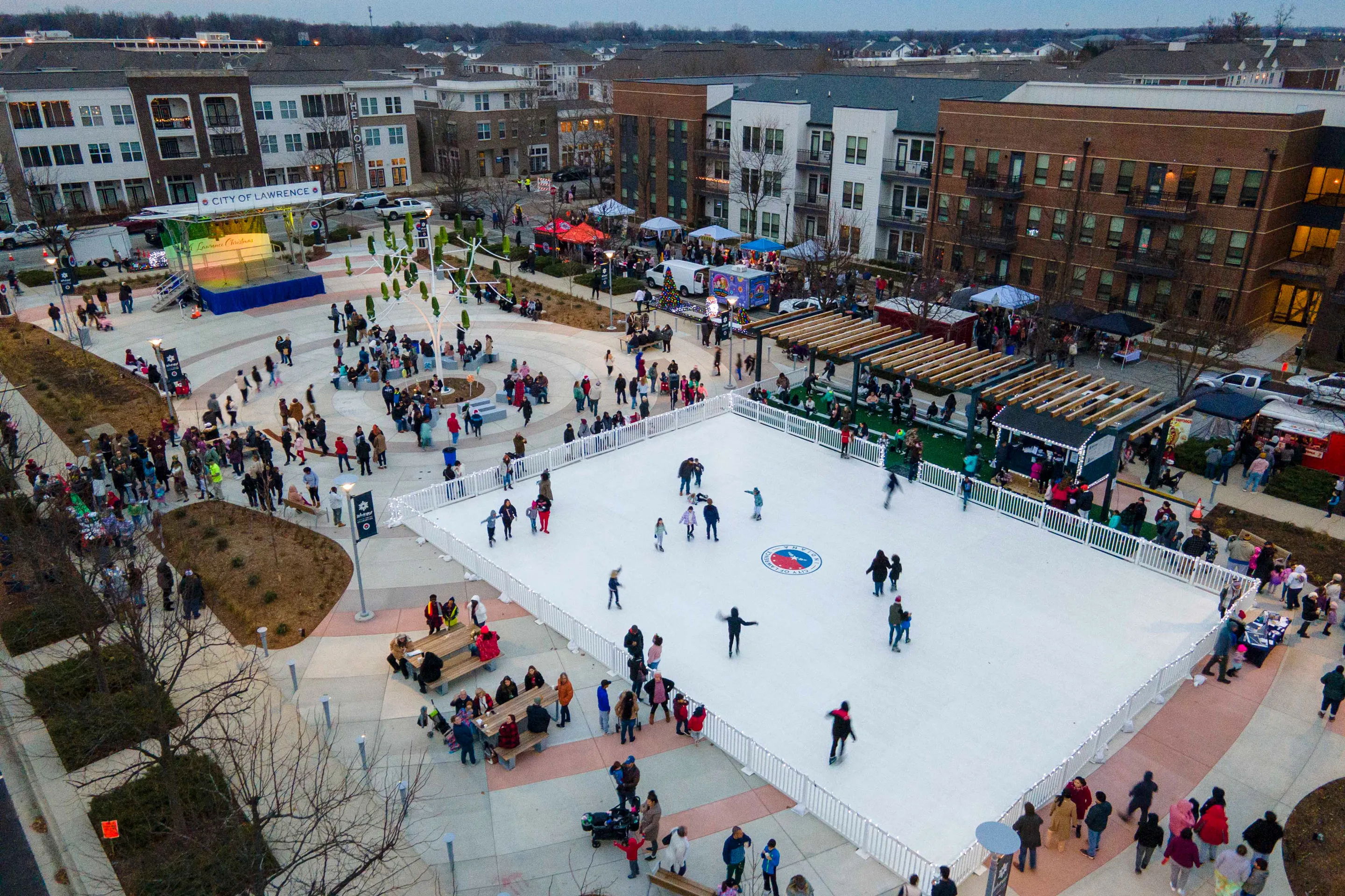 A lively outdoor ice skating rink filled with people enjoying winter festivities, surrounded by shops and colorful stalls.