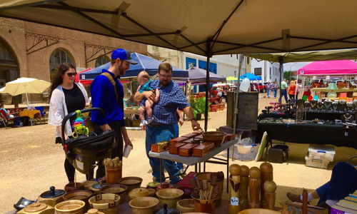 Outdoor market scene with vibrant handmade wooden items, a vendor, and families, evoking warmth and community.