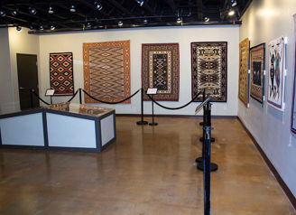 Art gallery displaying vibrant woven textiles and rugs, organized layout, warm lighting, inviting atmosphere for exploration.