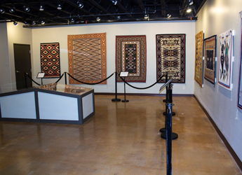 Art gallery displaying vibrant woven textiles and rugs, organized layout, warm lighting, inviting atmosphere for exploration.