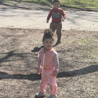 Two children walk joyfully in a sunny park, one in pink, the other in red, reflecting playful innocence and curiosity.